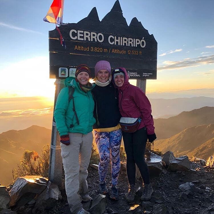 Three women in winter clothes stand in front of a sign for Cerro Chirripo with a sunrise shining in the background.
