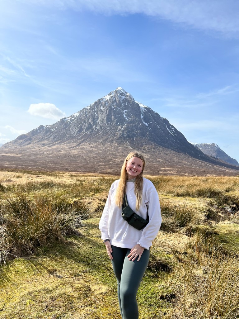 A girl with a shoulder backpack stands in front of a large mountain in the Scottish Highlands.