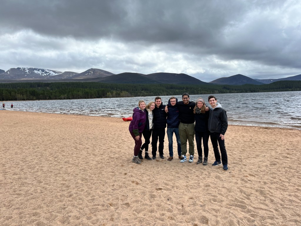Seven college students stand next to each other on a beach with the ocean and mountains in the background.