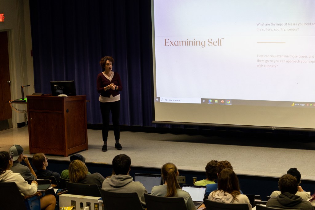 A woman stands on a stage in front of a projector lecturing to students who sit in seats in front of her.