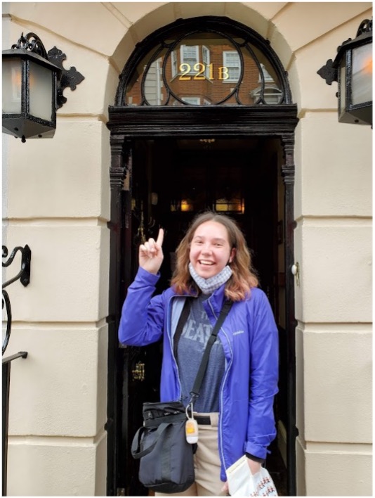 A girl stands in the entrance of a doorway pointing up at the number above the door, which reads 221.