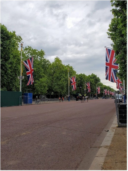 A wide brown street is lined with flagpoles where British flags are hung. Two horses mounted by guards stand in the middle of the street.