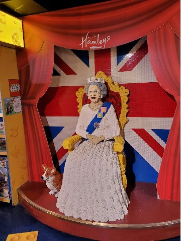 A lego statue of Queen Elizabeth sitting on her throne and her corgi sitting next to her sits in front of a large British flag.