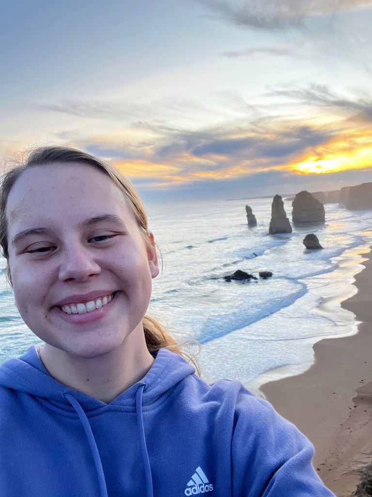 A girl in a blue hooded sweatshirt takes a selfie in front of a beach in Australia