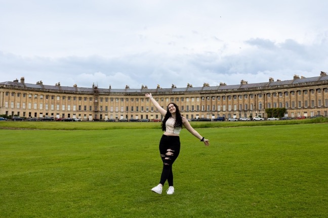A girl stands with her arms outstretched in front of the Royal Crescent in England.