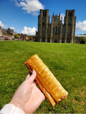 A sausage roll held up in front of Wells Cathedral in England