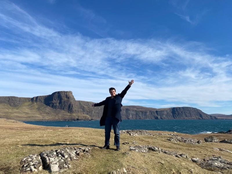 A man in a black coat stands with his arms outstretched in front of a mountain range and a large body of water.